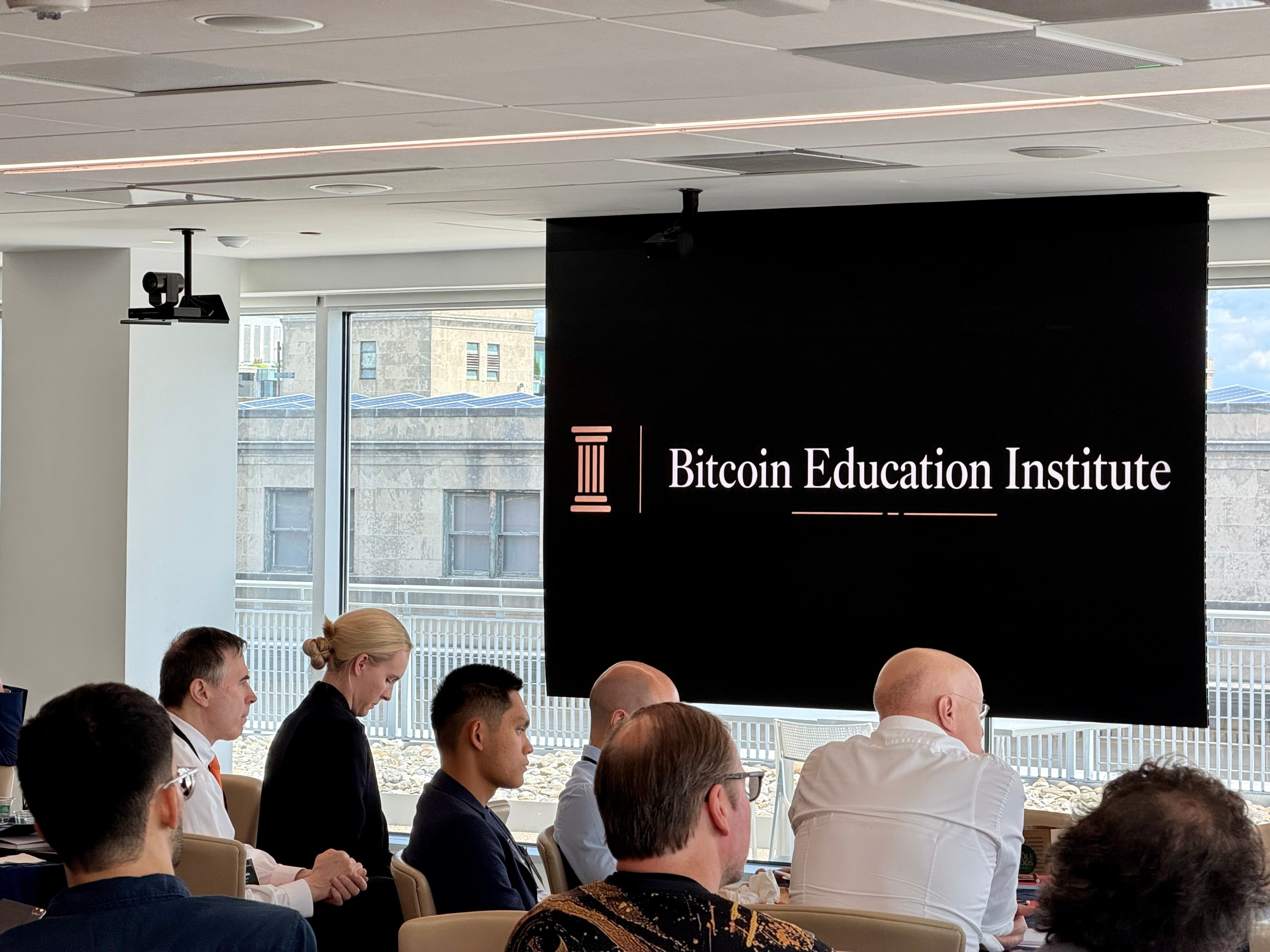 Attendees seated at the Bitcoin Education Institute conference facing a presentation screen carrying the new BEI column monogram and wordmark